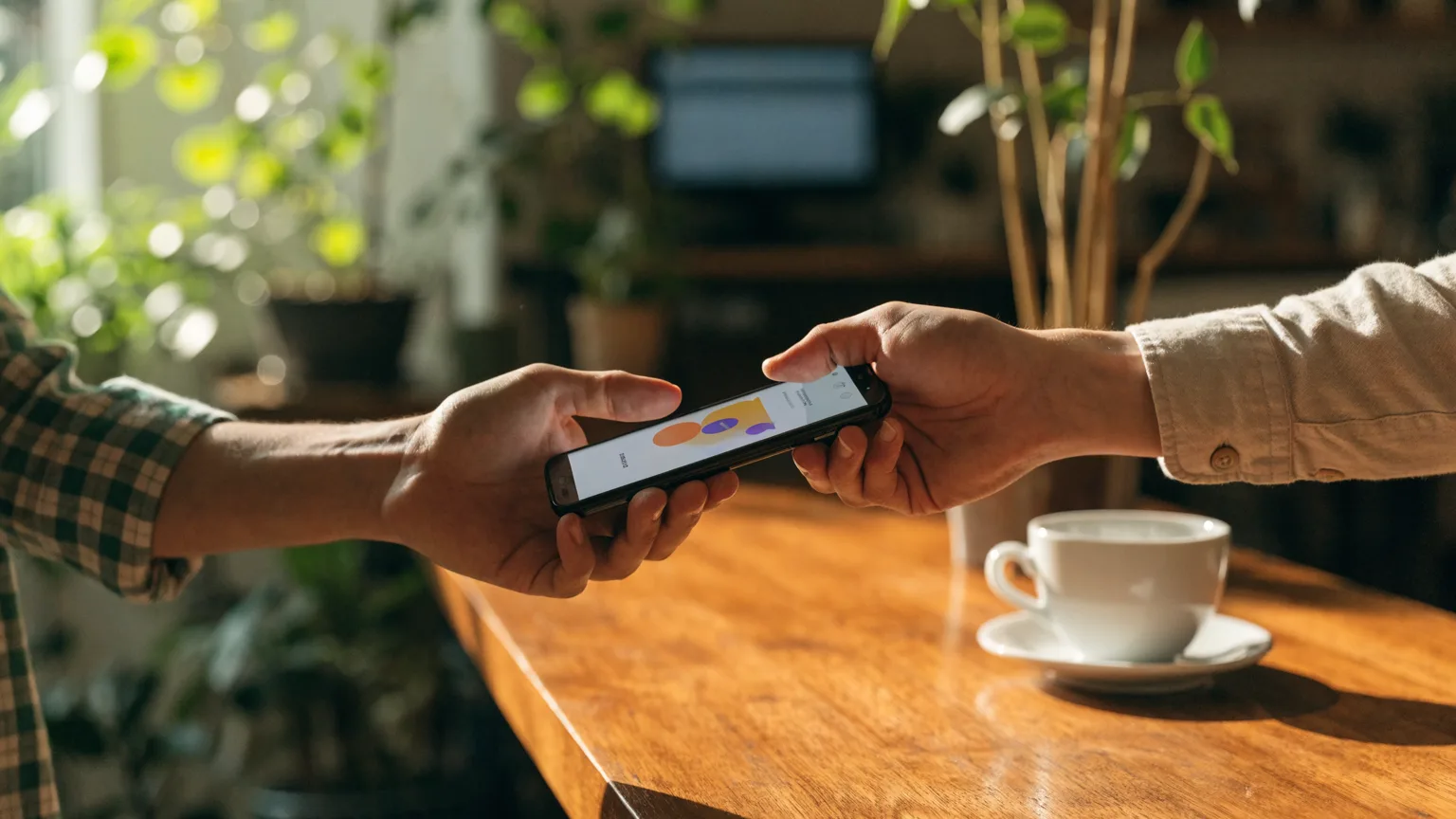 Mãos oferecendo um celular sobre o balcão de um pequeno negócio com uma mensagem de saudação na tela